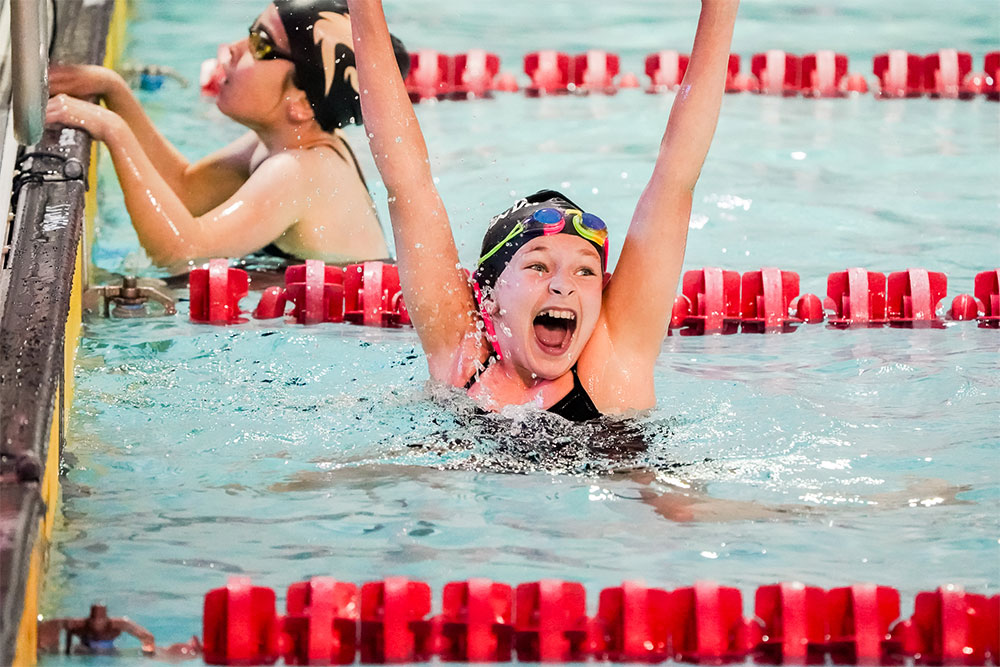 Young female swimmer with arms extended out of the pool, smiling widely with an excited expression